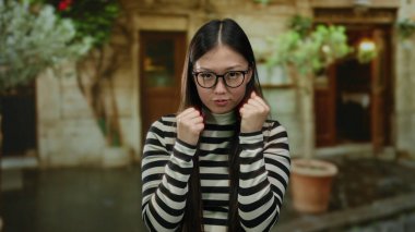 Şehir ortamında yumruklarını kaldırmış, gözlük takmış, çizgili gömlek giymiş şehir ortamında kendine güven ve güç sergileyen bir kadın..