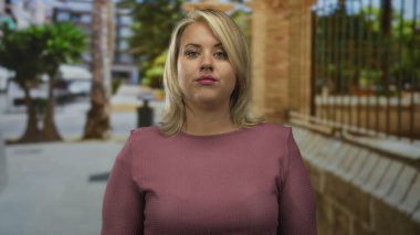 Elleri yanağında sarışın bir kadın işlek bir caddede demirden çitlerin ve palmiye ağaçlarının yanında dikiliyor..