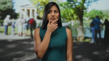 Parktaki genç Latin kadının, güneşli bir günde, etrafı bulanık insanlar ve yeşilliklerle çevrili dışarıda dururken bir fikri var..