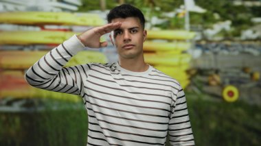 Sahilde askeriyeye selam veren genç bir adam, arka planda kanoları olan, çizgili gömlek giyen, kendinden emin görünen ve deniz tarafından bestelenmiş, açık hava macerasını ve saygıyı temsil eden biri..