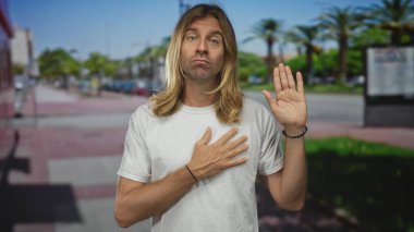 Beyaz tişörtlü, uzun sarı saçlı bir adam bir elini göğsünde tutuyor ve diğer elini güneşli bir sokak sahnesinde kaldırıyor..