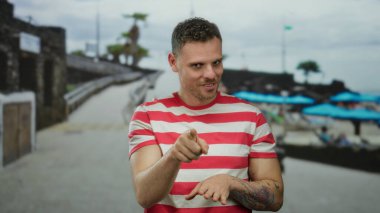 Kumsalda güneşli bir havada mavi şemsiyeler altında çizgili gömlek ve dövmeli yakışıklı bir adam.