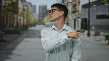 Gözlüklü genç İspanyol adam, yüksek binalarla çevrili, hareketli, güneşli şehir caddesini işaret ediyor..