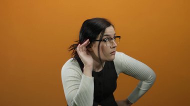 Gözlüklü ve esmer saçlı bir kadın turuncu bir arka plan önünde merakla ve dikkatle hareket ediyor..