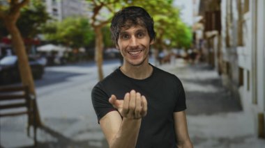 Gündelik siyah tişörtlü genç adam canlı şehir caddesinde kameraya doğru elle öpücük savuruyor; sevgi dolu karşılama romantizmi.
