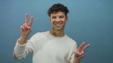 Mavi stüdyoda gülümseyen ve barış işareti yapan gözlüklü genç İspanyol adam iyimserlik.