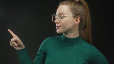 Stüdyoda parmağını kaldırmış, işaret parmağını kaldırmış, yana bakarken, gözlük ve yeşil süveter takan genç bir kadın..