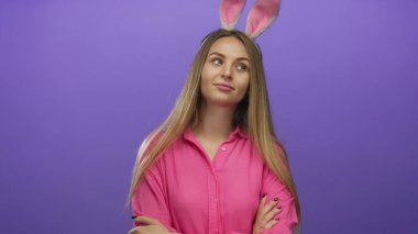 Tavşan kulaklı genç bir kadın, izole edilmiş mor bir arka plana karşı güvenle poz veriyor, canlı pembe bir gömlek giyiyor, neşeli ve neşeli bir paskalya temasını somutlaştırıyor..
