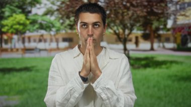 Beyaz tişörtlü genç adam yeşil bir parkta dikiliyor, düşünceli ve huzurlu görünüyor, elleri ağaçların altında bulanık arka planda dalgın dalgın hareketler yapıyor..