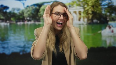 Neşeli bir şekilde gülümseyen bir kadın, güneşli yeşil bir parkta elleri ile yüzünü kapatarak, canlı bir arka plana karşı neşe sergiliyor..