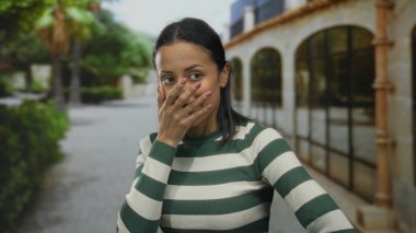 Çizgili süveter giyen genç İspanyol kadın, açık hava ortamında, kemerli pencereli, neşeli bir şekilde yüzünü kapatıyor..