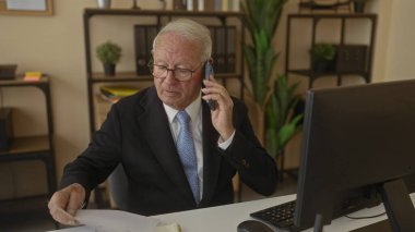 Takım elbiseli üst düzey bir adam ofis masasında bilgisayarlı ve kitaplıklı belgeleri incelerken iş görüşmesine katılmış..