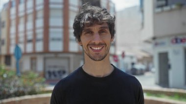 Kirli sakallı bir adam, güneşli şehir sokağındaki kameraya bina cephesiyle bakar..