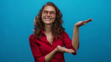 Kırmızı gömlekli kadın, mavi arka planda sunum yapar gibi ellerini uzatıyor, kıvırcık saçlar ve gözlüklerle gülümsüyor, izole ortamda pozitiflik tasvir ediyor..