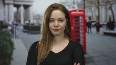 Genç kadın, işlek şehir caddesinde kırmızı telefon kulübesinin önünde bulanık yayalarla el ele tutuşuyor; inkar.