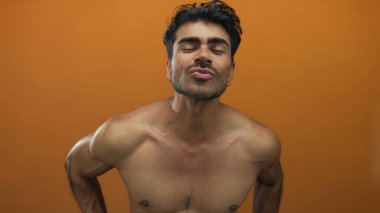 Turuncu stüdyo ortamında dudakları ve kaslı göğüsleriyle öpüşen bir adam rahatlatılmış kafasını eğip gülümsüyor..