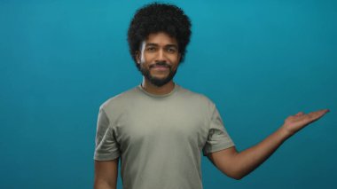 Mavi arka planda gülümseyen kıvırcık saçlı ve günlük giysileri olan bir adam izole edilmiş stüdyoda mutlu bir şekilde poz veriyor ve pozitif bir ifade sergiliyor.