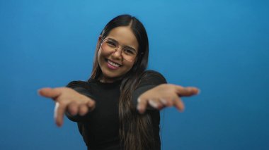 Mavi arka planda kollarını açmış gülümseyen genç bir kadın, gözlük takıyor ve siyah bir bluz giyiyor, izole edilmiş bir stüdyoda neşe ve açıklığın somutlaştırıcısı..