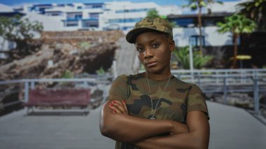 Kamuflaj üniformalı, çıplak kollu ve künyeli bir kadın binanın önünde şapka takıyor..