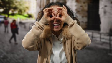 Kıvırcık saçlı, olgun bir İspanyol kadın güneşli bir caddede dürbünle el kol hareketi yapar..