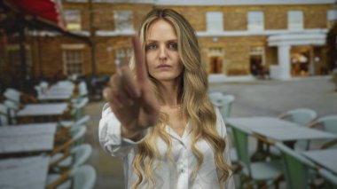 Uzun sarı saçlı bir kadın güneşli bir açık hava restoranının terasında arka planda boş masaları ve tuğla binaları olan kameraya doğru elini kaldırır..