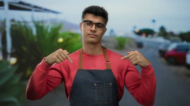 Gözlüklü genç adam önlük takıyor. Kenar mahalle caddesinde kendine güvenerek dikiliyor. Canlı bir şehir havasını kırmızı bir kazakla somutlaştırıyor..