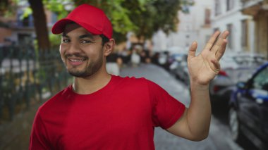 Kırmızı gömlekli ve şapkalı adam el sallıyor ve şehir sokaklarında coşkulu bir şekilde gülümsüyor; dostça selamlama.