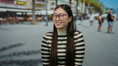 Şehir sokaklarında arka planda gülümseyen bir kadın çizgili gömlek giyen, parlak bir ifadeyle, uzun saç gözlüklü ve neşeli tavırlı bulanık yayalar.