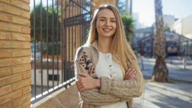 Güneşli bir günde şehrin arka planında ve yakınındaki tuğla duvarda çapraz kollarla güvenle gülümseyen bir kadın..