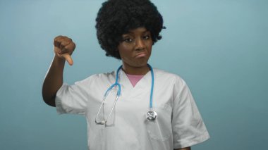 Steteskoplu beyaz önlüklü kadın doktor mavi stüdyoda baş parmağını kaldırıyor..
