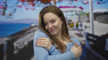 Genç kadın, açık mavi süveter içinde güneşli deniz kenarındaki gezinti alanında gözleri kapalı olarak kendine sarılıyor. Huzur..