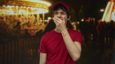 Kırmızı gömlekli genç adam, geceleri kalabalık caddede parlak fuar ışıklarının altında eli ile burnunu çimdikler; iğrenme.