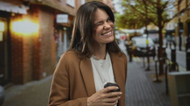 Şehir merkezindeki bir sokakta gülümseyen ve kahve tutan genç İspanyol kadın kalabalık bir kentsel alanda canlı ve neşeli bir atmosfer yakalıyor..