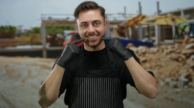 Sakallı, gülümseyen ve tebessümünü gösteren genç bir adam önlük ve eldiven takmış bir inşaat alanında arka planda bitmemiş bir evi olan..