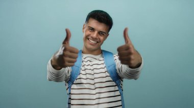 Mavi bir arkaplan önünde başparmağıyla gülümseyen genç adam, çizgili bir gömlek ve sırt çantası giyiyor, pozitifliğini ve güvenini ifade ediyor..