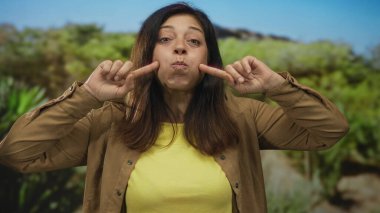 Kadın geniş gülümseme ile yeşil yapraklar ile güneşli orman sahnesinde iki işaret parmağı ile yanaklarını çimdikler;.