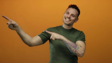 Genç adam gülümsüyor ve izole edilmiş turuncu bir arka planda dövmeli bir kolla pozitif ve çekici bir tavır sergiliyor..