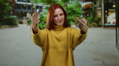 Şehir sokağında orta parmaklı bir kadın, sarı bir kazak giyiyor, kızıl saçlı, kendine güvenen bir ifade çiziyor, şehir arka planında bulanıklık var, davranışlarını vurguluyor.