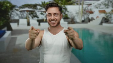 Otelin bilardo salonundaki kameraya gülümseyen sakallı genç adam, beyaz atlet giyiyor, el hareketi yapıyor, güneşli gökyüzünün altında açık hava otelinin tadını çıkarıyor..