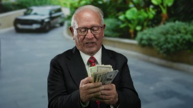 Kıdemli işadamı, arka planda lüks bir arabasıyla bize dışarıda dolar sayıyor, modern bir şehir ortamında zenginlik ve başarı sergiliyor..