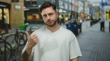 Beyaz tişörtlü, sakallı genç adam şehir caddesinde başparmak kaldırıyor. Etrafı dükkan ve bisikletlerle çevrili..
