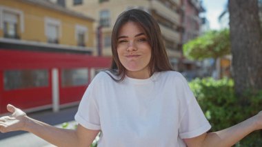 Gündelik beyaz gömlekli bir kadın arka planda yeşillik ve kırmızı binalar olan güneşli bir şehir sokağında kafa karışıklığı içinde açık havada şaşkın bir ifadeyi canlandırıyor..
