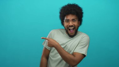 Gülümseyen Afro-Amerikan gözlüklü adam heyecan ve merak dolu bir ifadeyle canlı yeşil bir arkaplanı işaret ediyor..