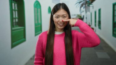 Bir şehir caddesinde pembe bir kazak giymiş, açık havada dururken burnunu gösteren kadın; şehir zemininde çekici ve neşeli bir anı yakalayan yeşil pencere çerçeveleri yer alıyor..