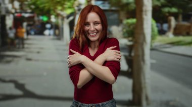 Şehir caddesinde gülümseyen, kendini kucaklayan, kızıl saçlı, şık kırmızı bir süveter giyen, bulanık arka planda mutlu görünen bir kadın..