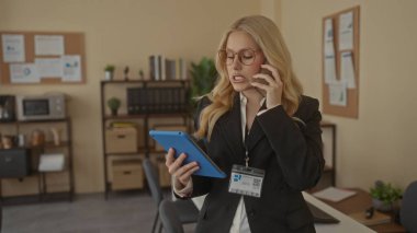 Gözlüklü genç sarışın kadın telefonda konuşarak ve modern ofis ortamında tablet kullanarak iş ve teknoloji etkileşimi sergiliyor..