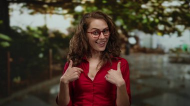 Kırmızı gözlüklü genç bir kadın parkın arka planında kırmızı tişört giymiş ve güneşli bir günde güvenle gülümsüyor.