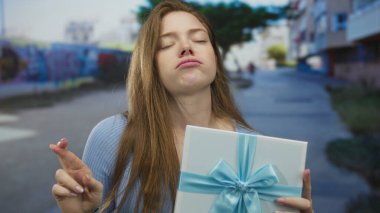 Beyaz bir kadın mavi hediye kutusunu tutarken kaşlarını kaldırarak yüzünü buruşturuyor..