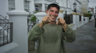 Yeşil kazaklı İspanyol adam kamerayı işaret ediyor ve şehir caddesinde yumruklarla yüzünü kapatıyor..