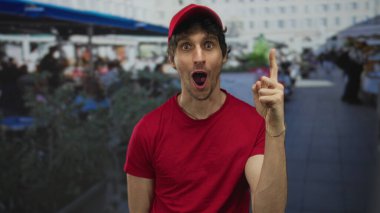 Kırmızı şapkalı ve kırmızı gömlekli adam şehir sokağının yanındaki kalabalık restoran terasında parmağıyla gökyüzünü işaret ediyor..